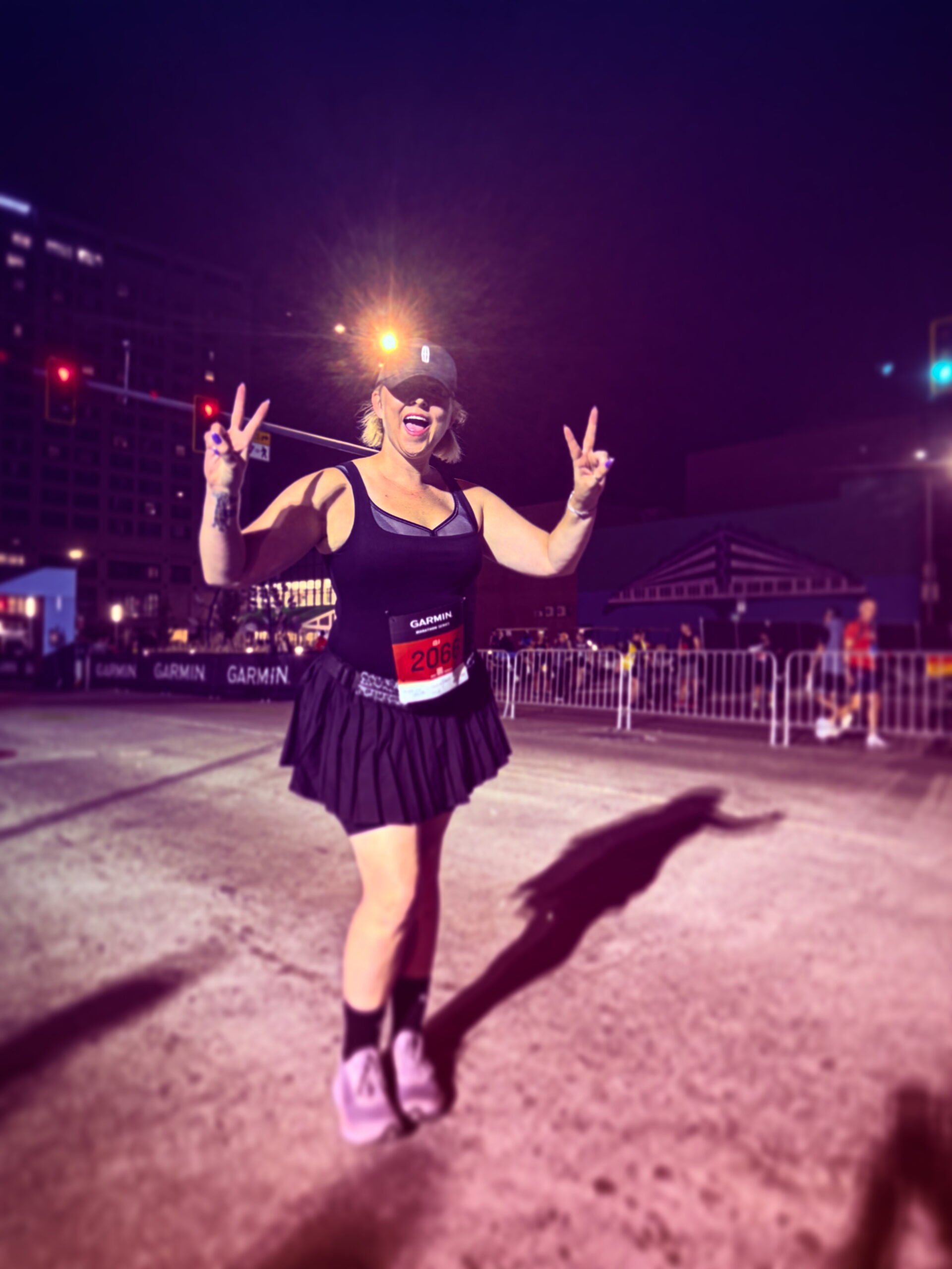 Kelsey in a black running skirt, tank top, and visor, holds up two peace signs on a city street at night. She is wearing a race bib and appears ready for a race.